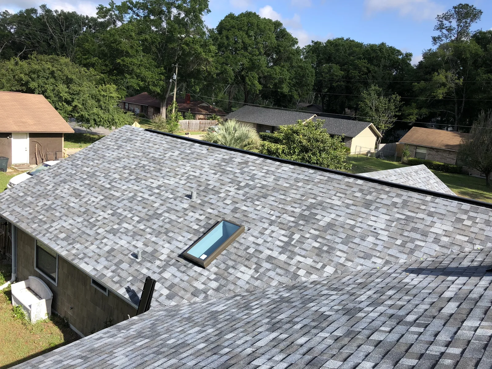 A gray shingle roof with a skylight over a single-story house, neighboring houses and tall trees behind in Holladay, UT