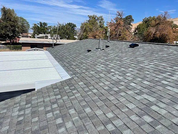 Gray shingle roof edge with roof vents and a low white flat section, mature trees and blue sky behind in Millcreek, UT